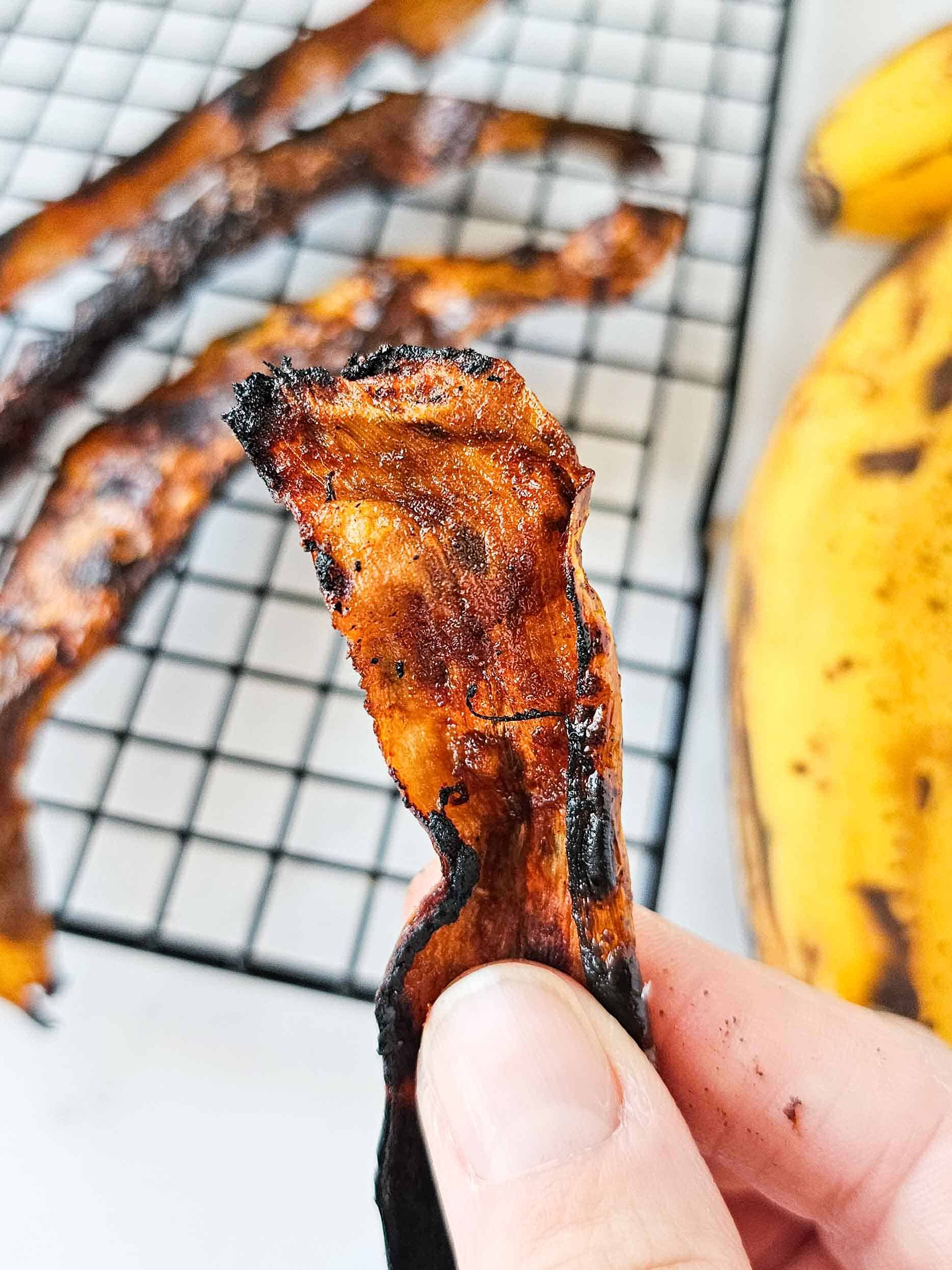 hand holding a slice of bacon with banana in the background